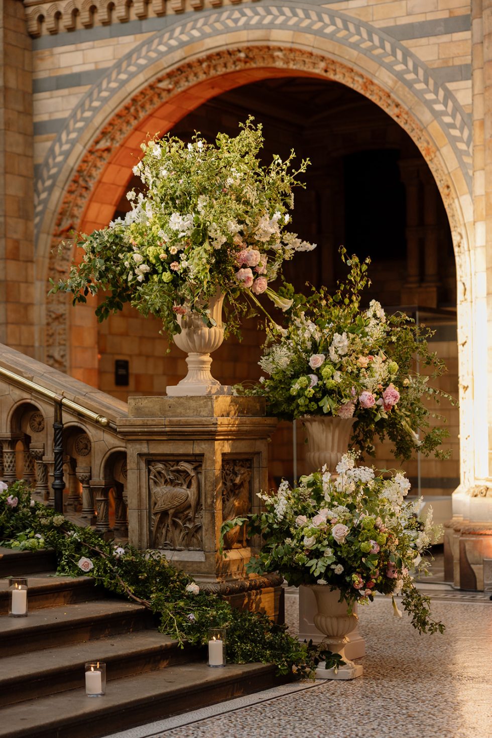 Luxury London wedding floral installations on the staircase of Hintze Hall at the Natural History Museum, featuring elegant urn arrangements of white, blush pink and green flowers with trailing greenery, styled as part of a luxury wedding ceremony and reception design.